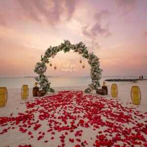 Romantic beach proposal setup in the Maldives at sunset, with a floral arch, hanging lanterns, and rose petals leading to a ‘Will you marry me?’ sign by the ocean.