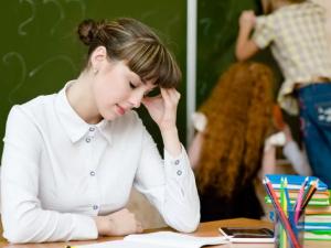 Teacher sitting at desk looking overwhelmed in classroom while students work in background