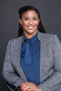 Angel G. Henry, International TEDx Speaker and Organizational Development Consultant, in a royal blue blazer with a navy shirt with side bow, smiling confidently against a dark grey background.