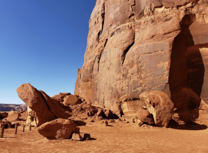 Tall sandstone rock face with weathered textures and faint markings, surrounded by scattered boulders on desert ground.