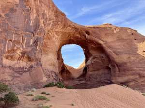 Wide view of a large sandstone arch with an opening framing the sky, surrounded by sand and sparse vegetation.