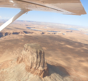 View from a small aircraft wing overlooking desert terrain and Lake Powell in the distance near the Arizona-Utah border