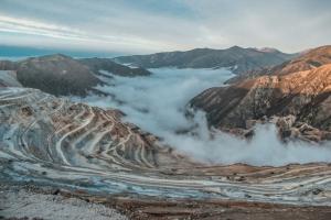 Vista amplia de una mina a cielo abierto en una región montañosa, con cortes en forma de escalones formando caminos sinuosos que descienden por la ladera; en el centro de la imagen, una capa densa de nubes o niebla cubre el valle, contrastando con los ton