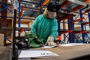 A student in welding safety gear, including gloves, safety glasses, and a raised welding hood, examines printed plans in an industrial training facility.