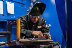 A student wearing a welding hood and safety glasses works closely to align small metal pieces at a workstation, focusing on precise alignment in a blue welding booth..