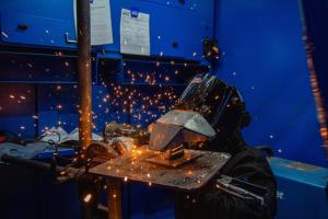A welding competitor wearing a protective welding hood performs an active weld on a small model, with sparks flying in a blue welding booth during the Project MFG Maritime Welding Competition.