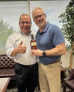 Two white men standing side by side in an office. One is holding up a bottle of liquor in honor of having just closed on the acquisition. Both men are smiling.
