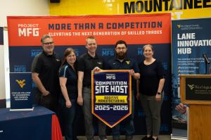 Five adults stand in front of a Project MFG backdrop at West Virginia University, holding a banner that reads “Competition Site Host – Additive Manufacturing 2025–2026,” with a podium and signage for the Lane Innovation Hub visible nearby.