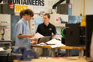 A Project MFG employee in a black polo stands beside two student competitors in blue uniforms as they review printed plans and a laptop in a manufacturing lab filled with CNC machines and equipment.