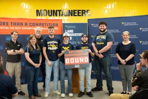 Members of West Virginia University’s Engineering Technology (ETEC) team stand with event staff holding a “National Championship Bound” sign after earning first place in the Project MFG Additive Manufacturing competition, with university and Project MFG b