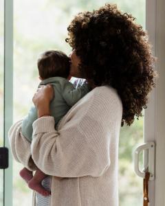 Mother holding her baby near a window in a soft nursery setting, representing everyday caregiving for babies.