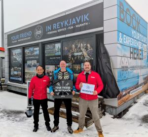 Three team members standing outside the Your Friend In Reykjavik office in Reykjavík, Iceland — two wearing red Whisper-branded jackets and one holding a Your Friend In Reykjavik sign, representing the new Whisper Iceland partnership.