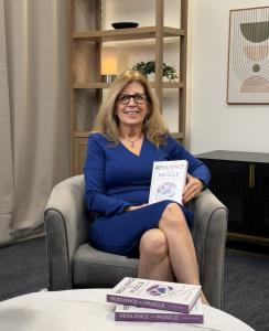 Laurie Carey sitting in a chair holding her book Resilience Is a Muscle, with additional copies on a table, representing her work supporting We Connect The Dots mission
