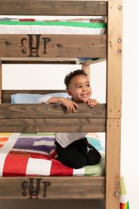 A young boy with curly hair sits on the lower bunk of a handmade wooden bunk bed, smiling and resting his chin on the bed rail. The letters "SHP" are carved into the bed frame. Colorful quilted bedding is visible on both bunks.