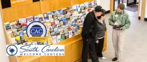 Three people at South Carolina Welcome Center, one staff with tablet assisting two visitors near travel brochures.
