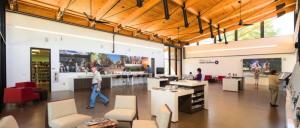 Bright, modern visitor center interior with high wooden ceilings, seating areas, display tables, large photo murals, and people exploring exhibits.