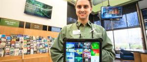 Person in uniform holding a tablet displaying a digital interface inside a visitor center with brochures and TV screens.