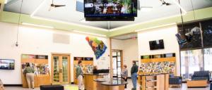 Interior of a visitor center with brochures, a large South Carolina map, seating, and people interacting at information desks.