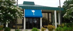 Entrance of a building with a green roof, blue awning featuring a white palmetto tree and crescent moon, surrounded by greenery and flowering trees.