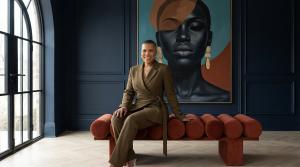Marki Lemons Ryhal, CSP, global keynote speaker, five-time Hall of Fame inductee, and founder of MenopauseOS, photographed in an olive green wrap blazer seated on a deep red velvet bench in a navy blue paneled room with a large-scale contemporary portrait
