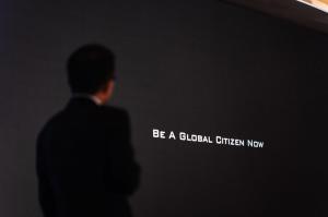 Silhouette of Henry Fan, CEO of Globevisa Group, from behind, looking at a large black screen with the white text "BE A GLOBAL CITIZEN NOW" at a Global Citizen Conference.