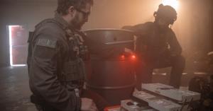 Two police tactical officers in full protective gear organise medical supplies from an open rugged case inside a smoke-filled industrial setting, with integrated red emergency lighting enhancing visibility in low-light conditions.