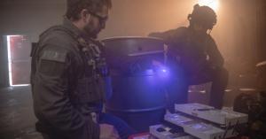 Two police tactical officers in full protective gear access organised medical supplies from an open rugged case inside a smoke-filled industrial environment, illuminated by blue task lighting to support visibility in low-light emergency conditions.