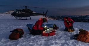 Mountain rescue team members in red protective gear administer medical assistance to a casualty on a snowy mountainside at dusk, using an organised rugged case for supplies, with a helicopter on standby in the background.