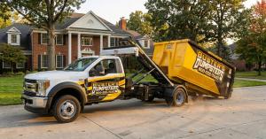 Buster's Dumpsters branded white hook-lift truck delivering a yellow roll-off dumpster to a residential driveway in Jackson MI for a home project
