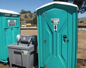 Two turquoise portable toilets mounted on a trailer parked on a residential street, featuring a large orange dinosaur decal on the left unit.