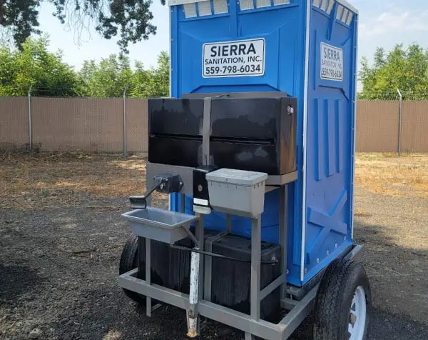 Close-up of a blue Sierra Sanitation portable toilet on a trailer, highlighting the exterior handwashing station with a sink and water reservoir.