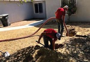 Two technicians in red shirts and caps performing a septic tank pumping and inspection service in a residential yard.