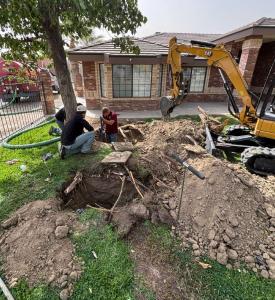 Plumbers using a mini excavator and shovels to dig a trench for sewer line repair in front of a brick house.