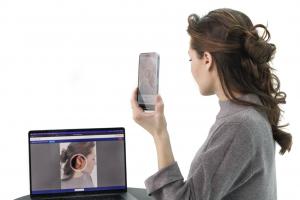 A woman holds a smartphone up to her ear while using an app to scan or measure it, with a laptop beside her displaying a live image of her ear with a highlighted target area.