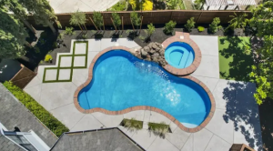 Aerial view of a modern backyard featuring a free-form pool, circular attached hot spa with rock waterfall, and large square concrete pavers set in artificial turf, with new privacy planting along a fence.