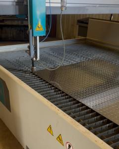 A CNC waterjet machine at Raw Welding cutting a sheet of industrial diamond plate metal. The teal cutting head is positioned over the metal on a large submerged grate, prepared for precision fabrication used in crane and rigging services.