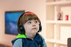 Young child wearing headphones and a blue and green zip-up jacket, looking upward thoughtfully in a home setting.