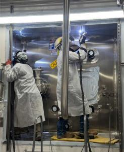 Two professional welders in white protective suits and welding helmets working on a large stainless steel industrial unit. One welder is focused on an upper corner seam with a visible welding arc, while the other inspects the side. This precision fabricat