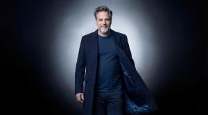 A professional portrait of singer and actor Gerónimo Rauch wearing a navy blue coat and t-shirt, looking at the camera against a dramatic studio background with a central light glow.