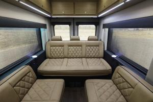 Interior view of the rear of a Waldoch StarLux camper van, showcasing tan diamond-stitched leather seating, including two side seats and a three-person rear sofa. The cabin features dark wood flooring, overhead wood cabinets with LED strip lighting, and l