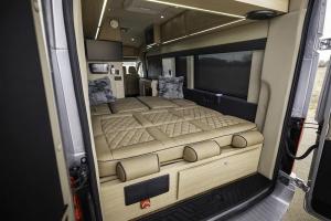 Interior view from the rear of a Waldoch StarLux camper van, showing the rear sofa converted into a large bed with tan diamond-stitched leather. The cabin features wood-grain cabinetry, a mounted Smart TV, and integrated LED ceiling lighting.