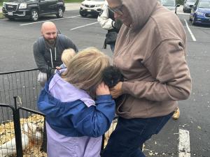 A young girl in a blue jacket presses her face into a small black fluffy chick being held by an adult in a tan hoodie. A smiling man and a toddler are visible in the background near a fenced animal enclosure.