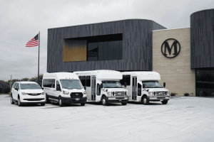 Four white commercial vehicles, including a minivan, a Ford Transit van and two shuttle buses, parked in front of Master's Transportation's modern Kansas City, Mo., headquarters building featuring the company's "M" logo.