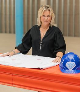 Pat Miller, the president of Blue Diamond Construction, is standing behind a red desk with blueprints and a blue hard hat laying on top.