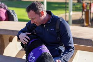 Guide Dogs of America - Veteran with his service dog laying across his lap, as he looks down with his arms wrapped around him in a moment of comfort..