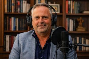 Author and interviewer Paul Rushworth-Brown recording a podcast interview with headphones and microphone in a home studio with bookshelves.