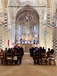 Interior view of a historic abbey church in Sezzadio, Italy, with attendees seated facing the altar during a commemorative Mass, beneath vaulted ceilings and a large religious fresco.