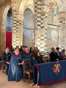Seated attendees , including individuals in ceremonial robes, listen during a Mass inside a historic abbey with stone columns and arched architecture in Sezzadio, Italy.
