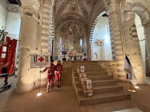 View of the altar inside a historic abbey church with clergy officiating a Mass, flanked by flags and Red Cross representatives standing beside a banner, beneath vaulted ceilings and frescoed walls.