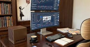 Kubb® Spectre Bronze compact workstation on a professional desk with dual monitors, keyboard and financial newspapers, showcasing its premium aluminum design.